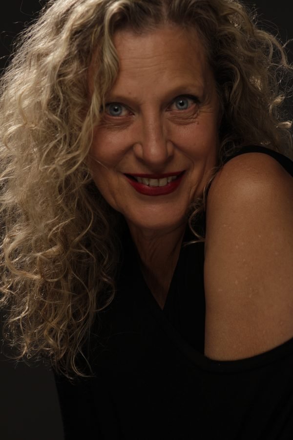 Retrato artístico de Mónica como Mona: primer plano con melena rizada suelta, labios rojos y mirada intensa a cámara, sobre fondo oscuro