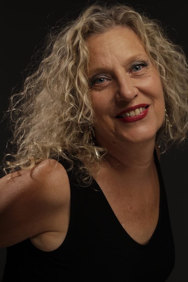 Retrato de estudio de Mónica Ibáñez Fernández de Lis: mujer de melena rizada y rubia, sonriendo a cámara con los labios pintados de rojo, sobre fondo oscuro